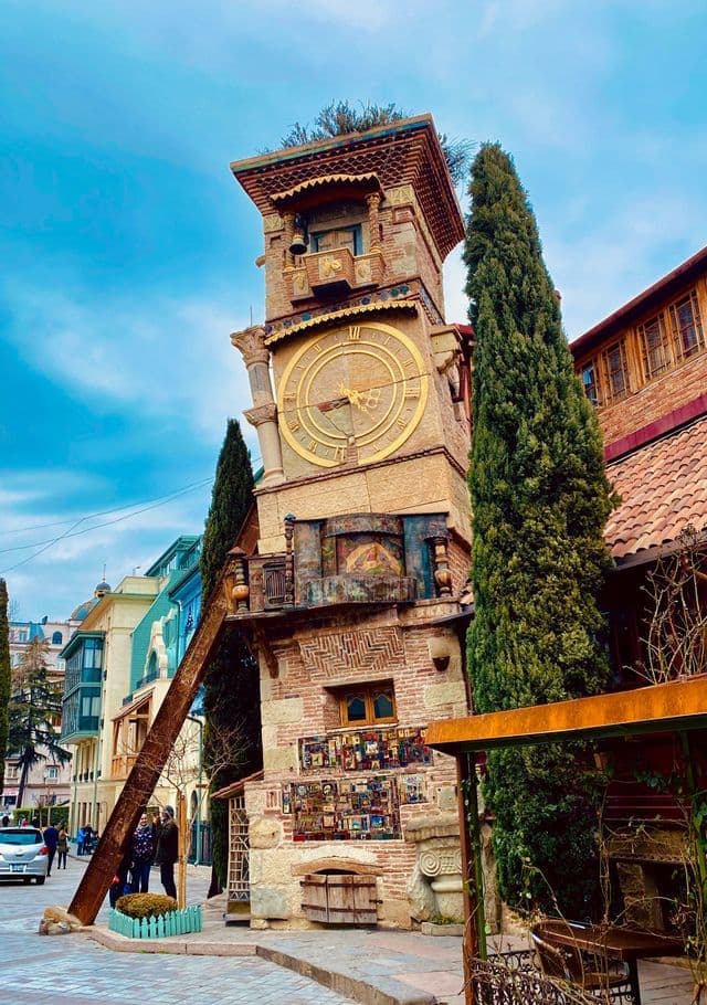 Una torre dell'orologio eclettica e pendente con un grande quadrante dorato si erge su una via cittadina accanto ad alti cipressi.