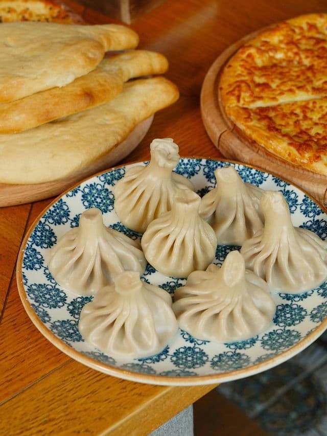 Un piatto di khinkali georgiani si trova su un tavolo di legno accanto a pane shoti e a un khachapuri al formaggio.