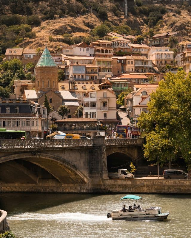 Un battello turistico solca un fiume, passando davanti a un ponte di pietra e a una città storica costruita su una collina, con una torre dal tetto verde.