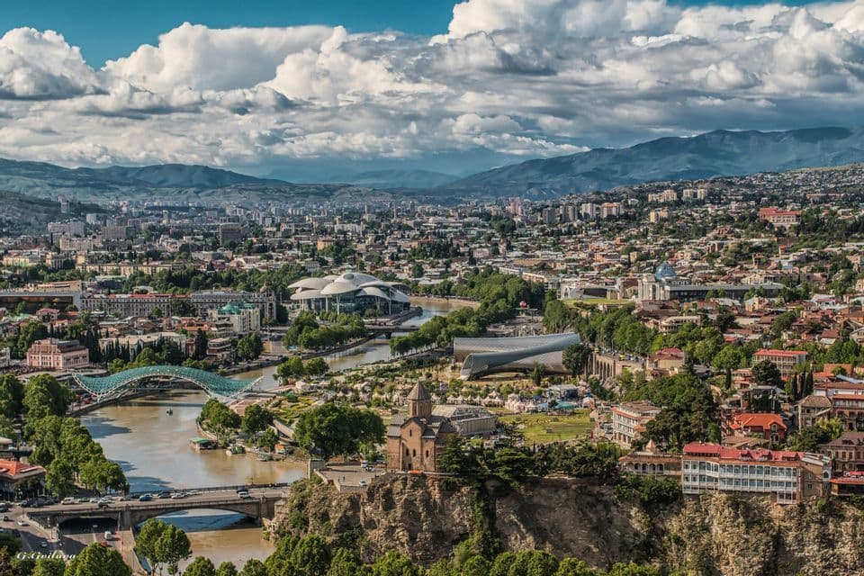 Una vista panoramica di una città con un fiume, architettura moderna e antica, e montagne lontane sotto un cielo nuvoloso.