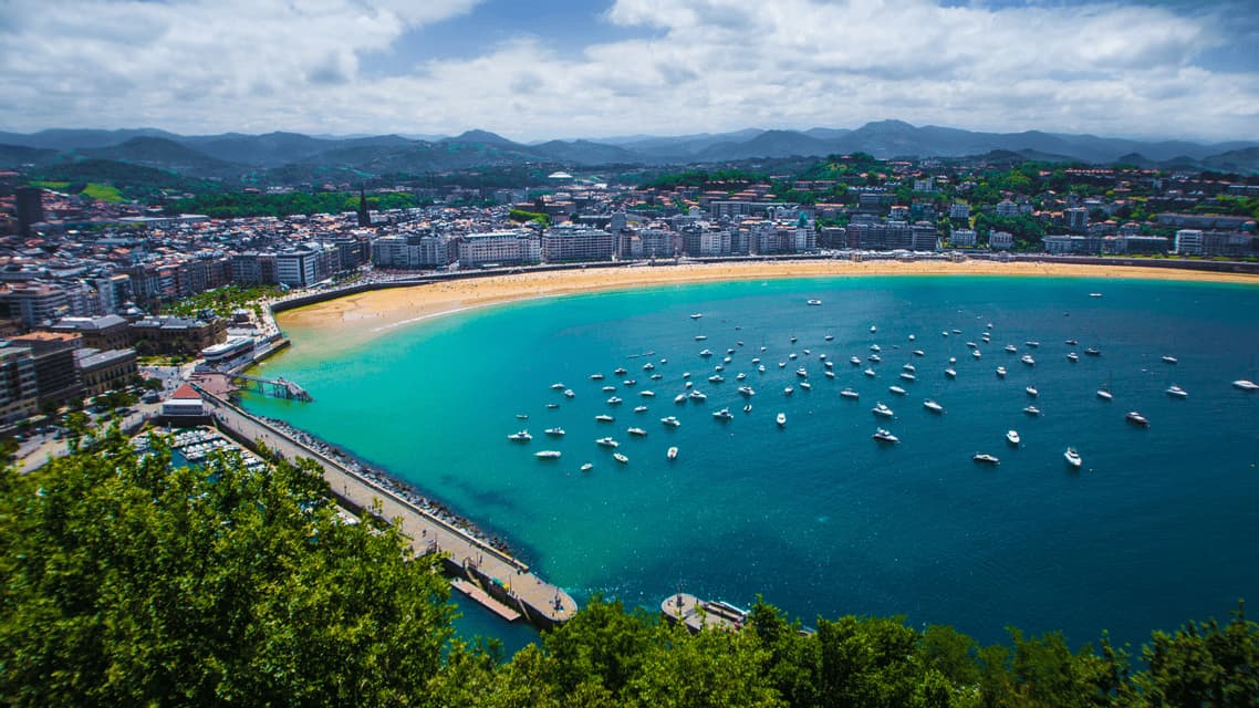 Una vista aerea di una città costiera con una spiaggia sabbiosa che si curva intorno a una baia piena di molte barche ancorate.