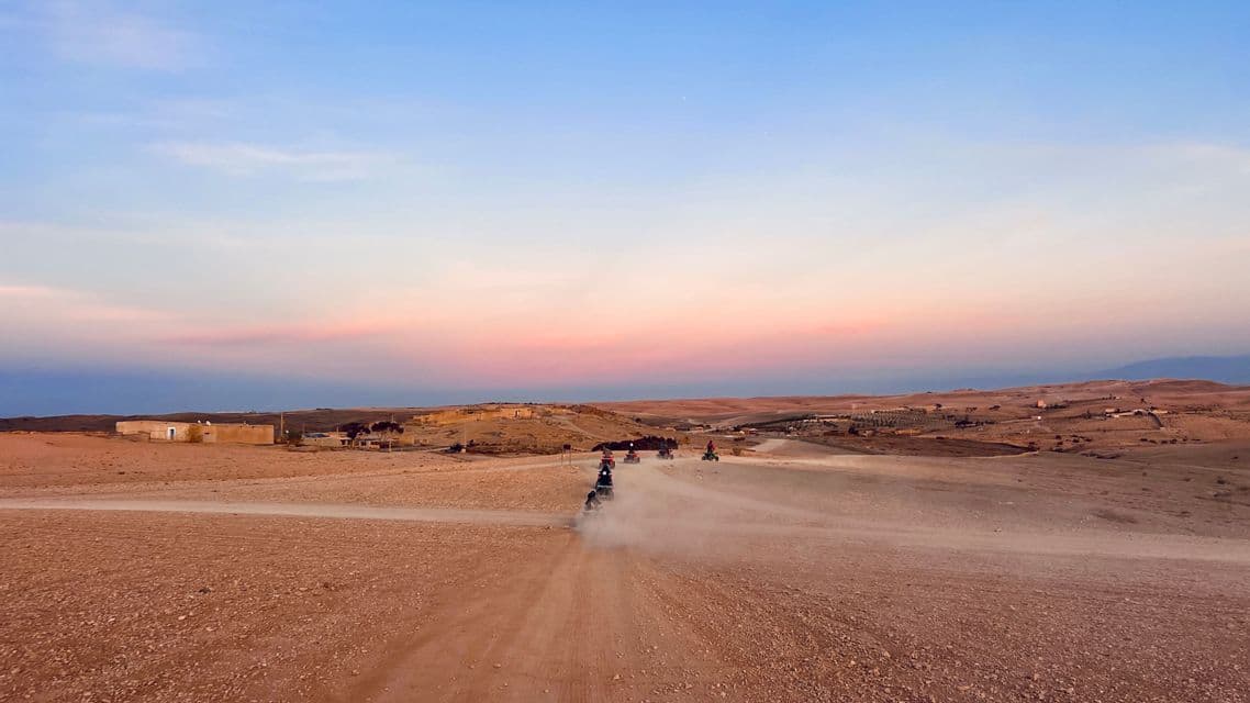 A WeRoad group trip riding quad bikes on a dusty track through a desert landscape during sunset.