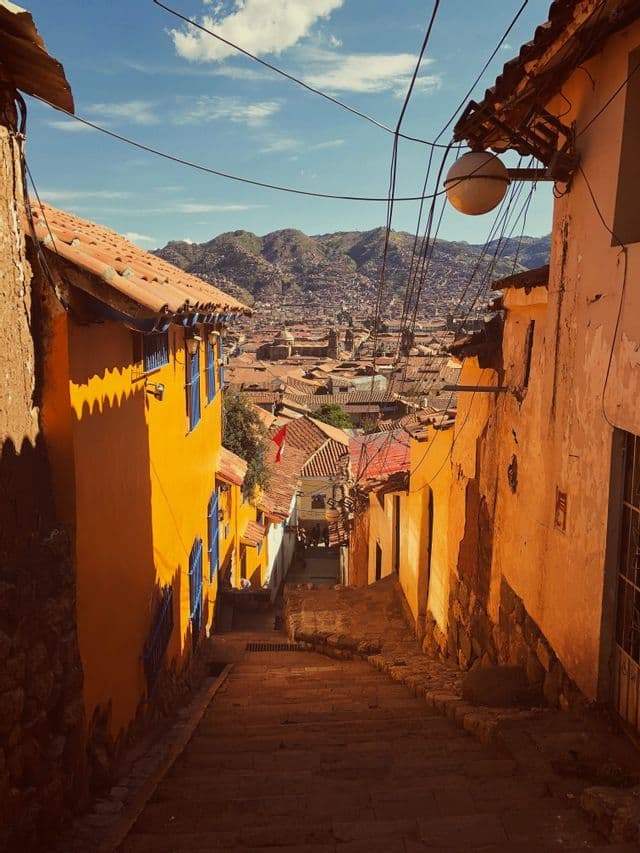 Un callejón de piedra empinado y estrecho con edificios coloridos domina una ciudad con tejados de terracota anidada entre montañas verdes.