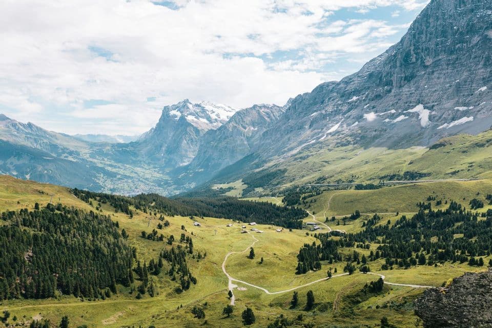 Una vista dall'alto di una valle montana verdeggiante con sentieri tortuosi e case sparse, sotto grandi cime innevate.