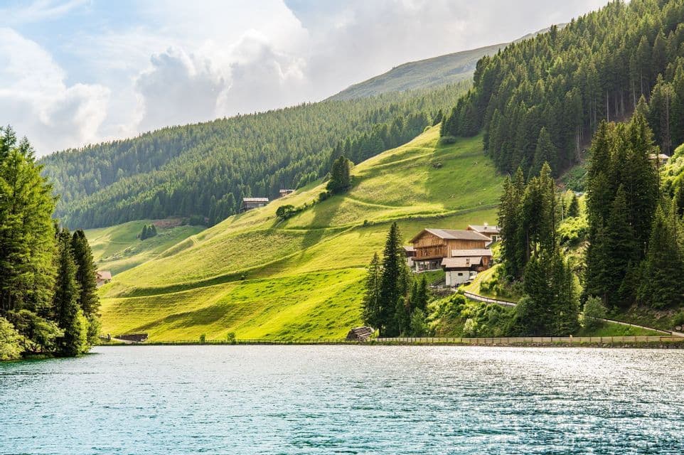 Un lago tranquillo si adagia ai piedi di una montagna verde e rigogliosa, punteggiata di case in legno e ricoperta di pini.
