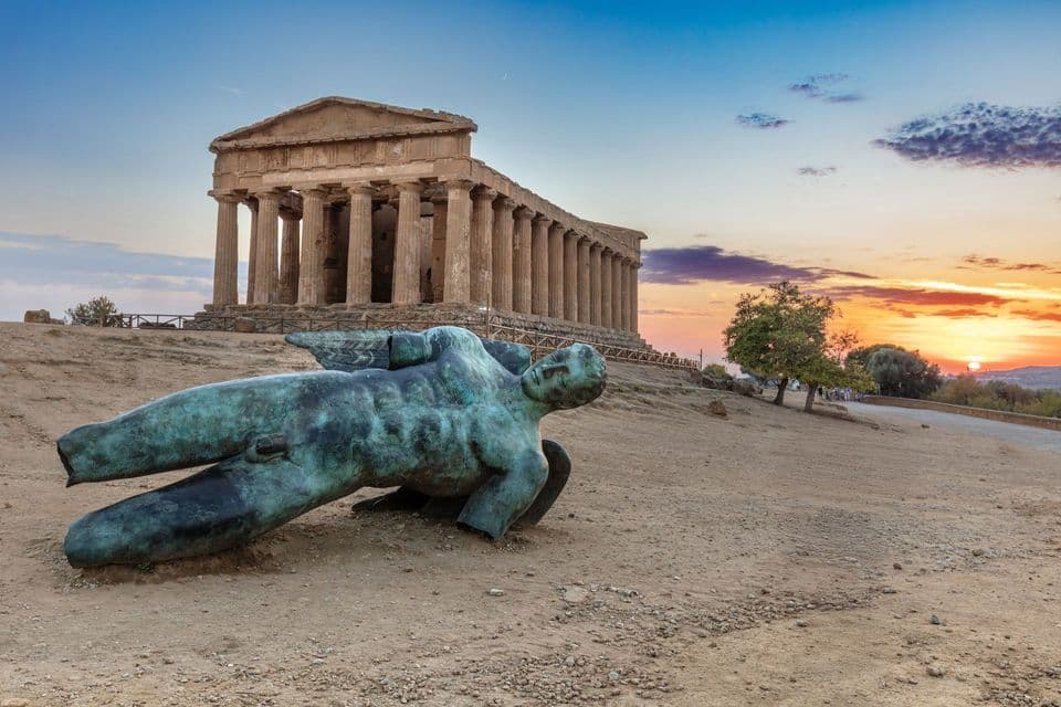 A bronze statue of a fallen winged figure lies on a dusty hill with an ancient Greek temple in the background at sunset.