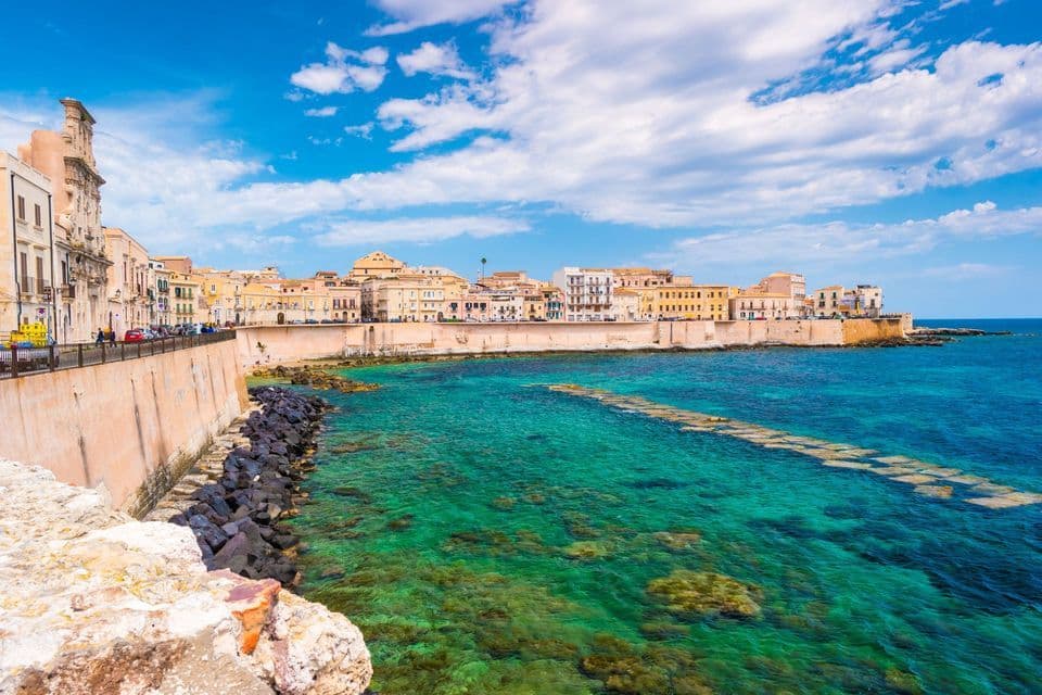 A historic coastal town with a seawall bordering clear, turquoise water under a blue, cloudy sky.