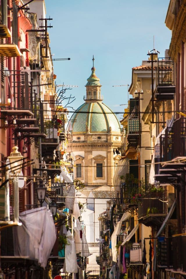 A green-tiled dome of a church is visible at the end of a narrow street lined with colourful buildings and hanging laundry.