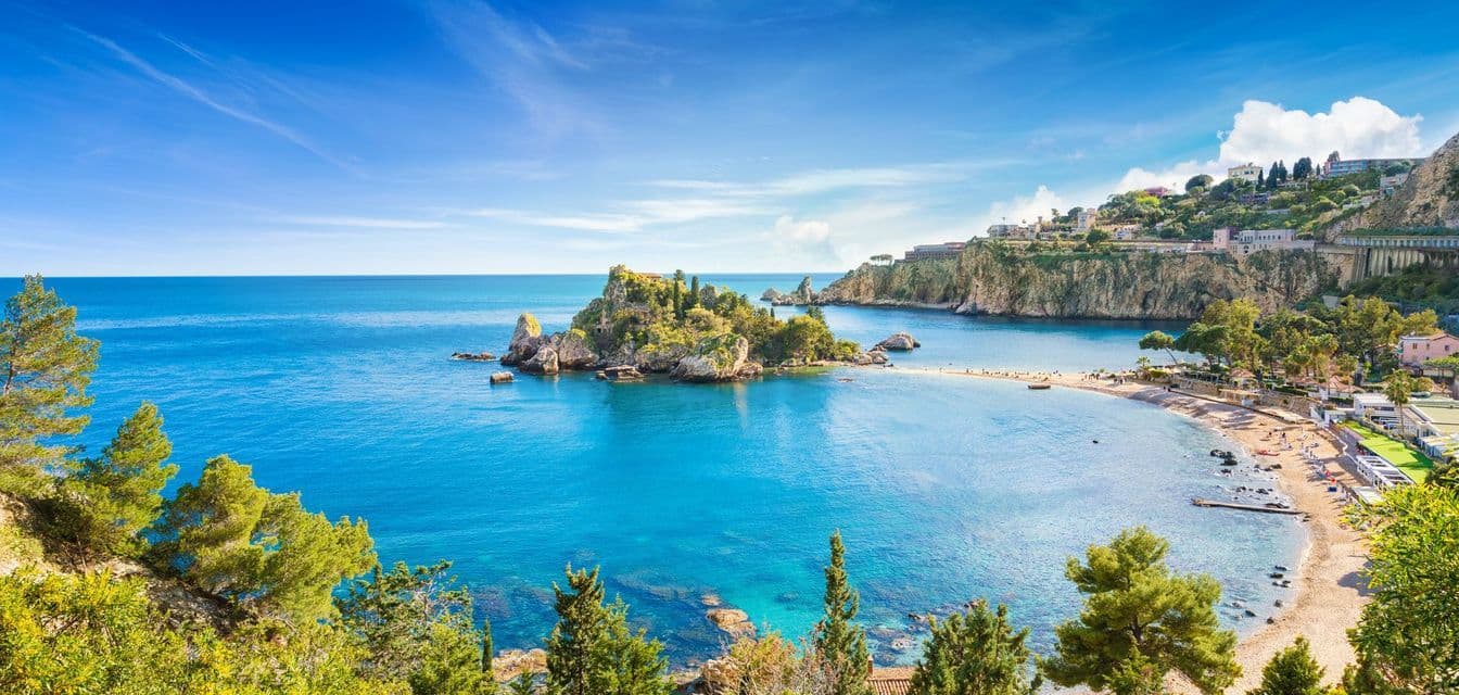 A panoramic view of a bay with a small, green island connected by a sandbar to a beach along a cliffside coast.