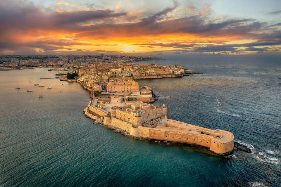 An aerial view of a large stone fortress on a peninsula with a coastal city behind it, all under a vibrant sunset sky.