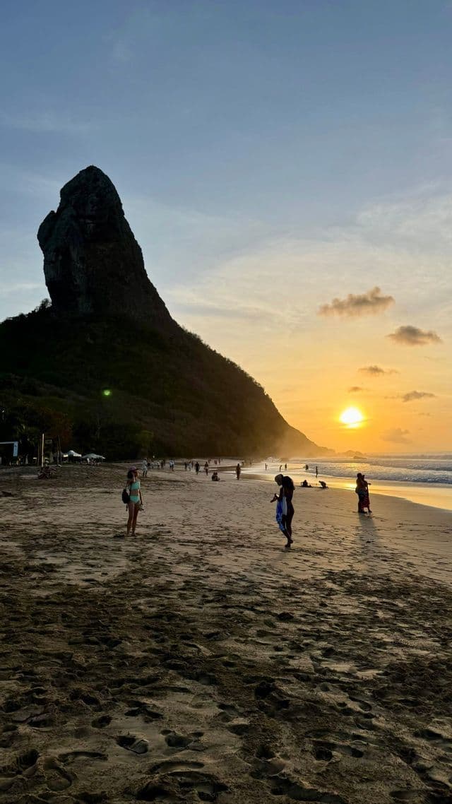 Persone passeggiano su una spiaggia sabbiosa durante un tramonto dorato, con una grande formazione rocciosa scura sullo sfondo.
