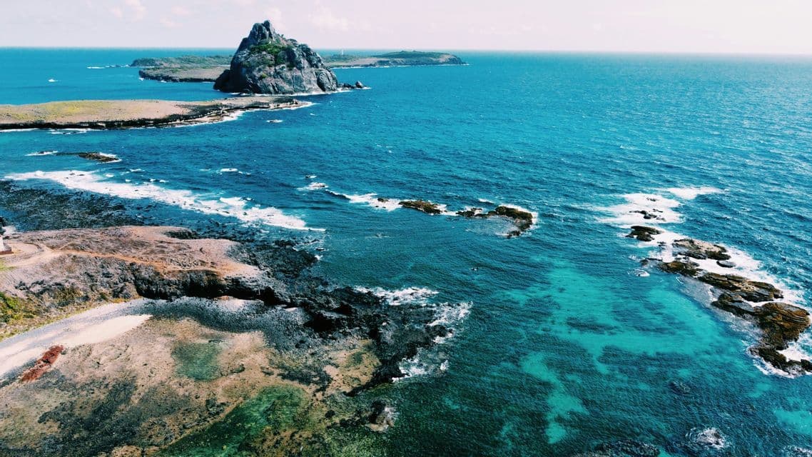 Vista aerea di una costa frastagliata dove l'oceano turchese e blu intenso incontra la riva rocciosa, con isole all'orizzonte.