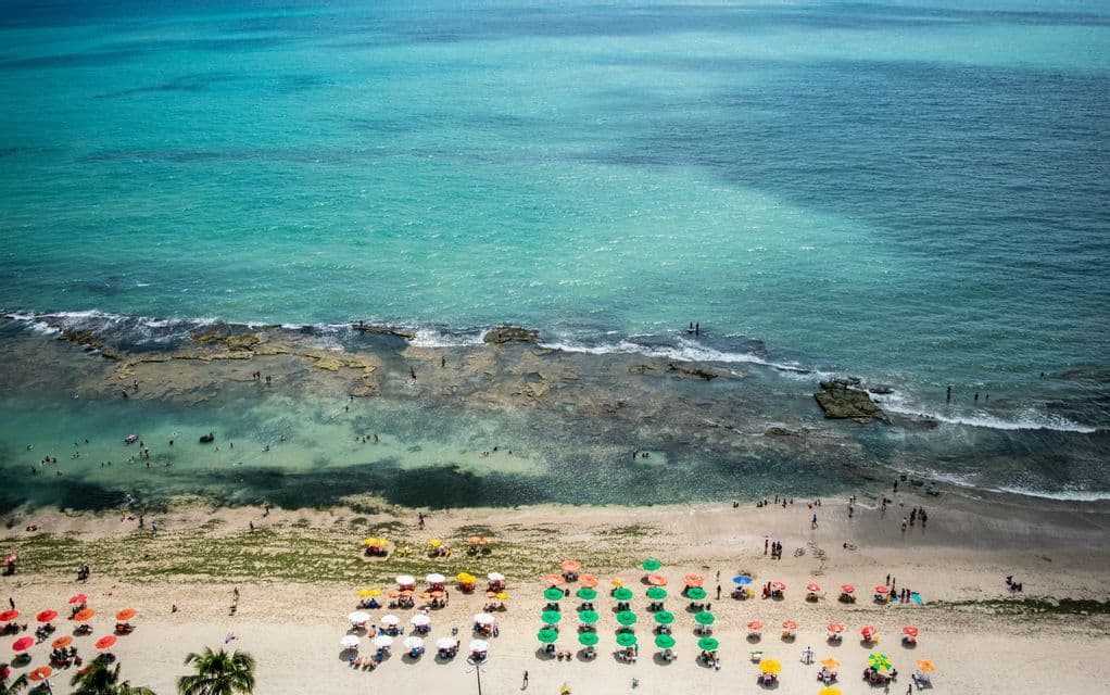 Una veduta aerea di una spiaggia affollata, con file di ombrelloni colorati sulla sabbia e persone che nuotano nel mare turchese.
