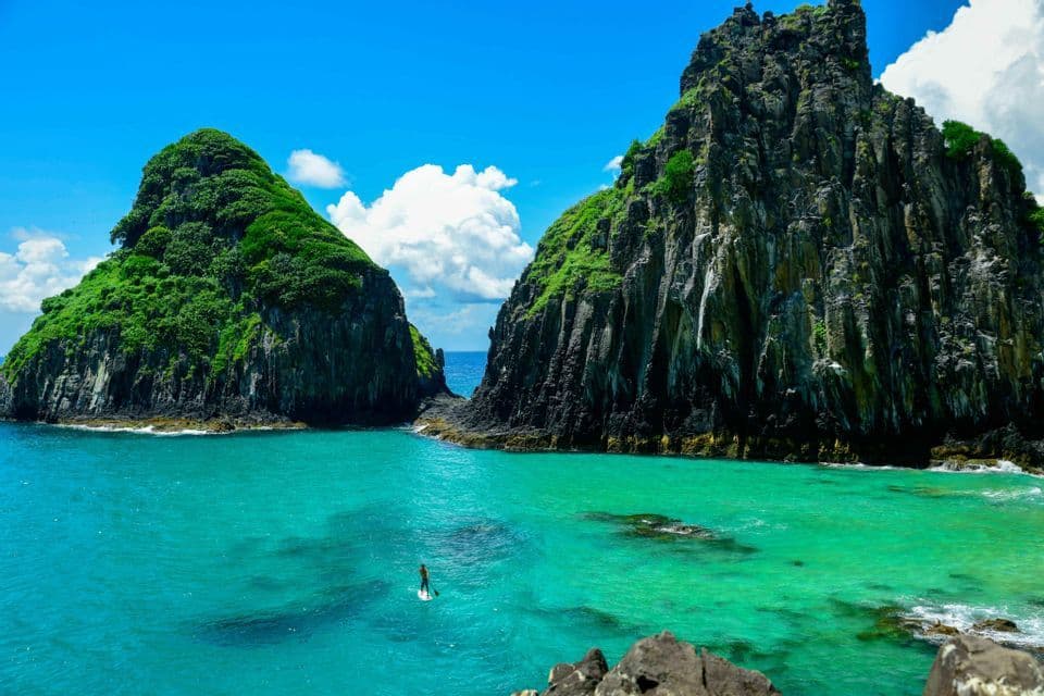 Una persona su un paddleboard naviga in acque limpide e turchesi tra due grandi e ripide formazioni rocciose sormontate da vegetazione verde.
