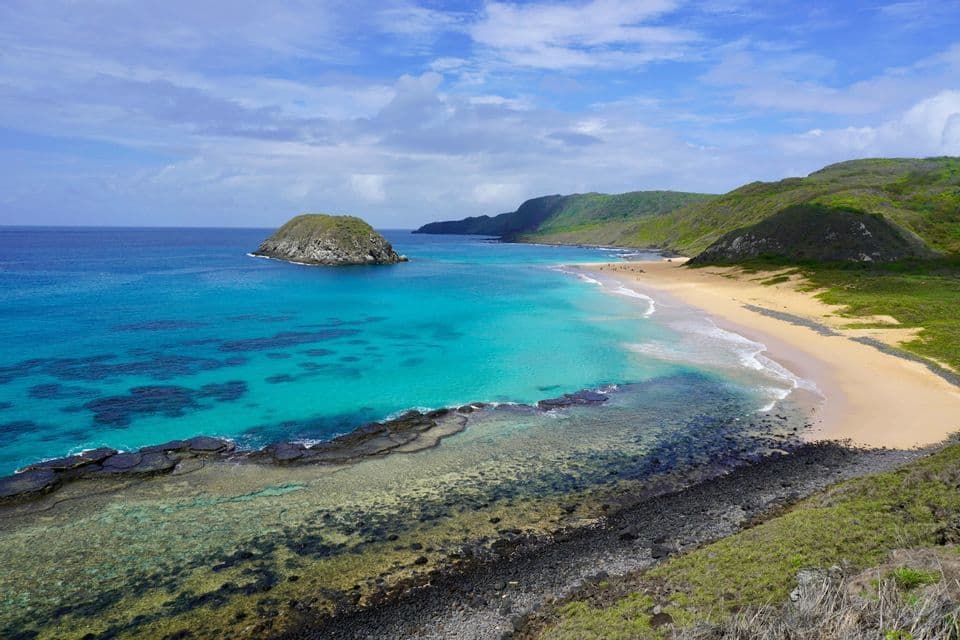 Una vista dall'alto di una baia costiera con acqua turchese limpida, una spiaggia sabbiosa e colline verdi sotto un cielo parzialmente nuvoloso.
