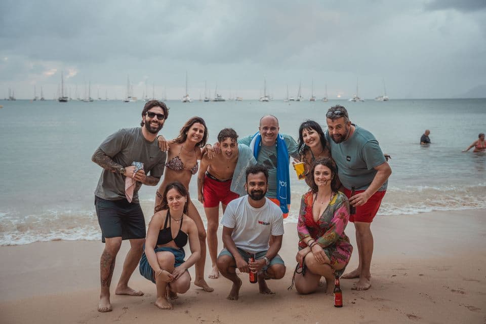 Nove persone di un gruppo WeRoad sorridono in una foto su una spiaggia sabbiosa, con il mare e barche a vela sullo sfondo.