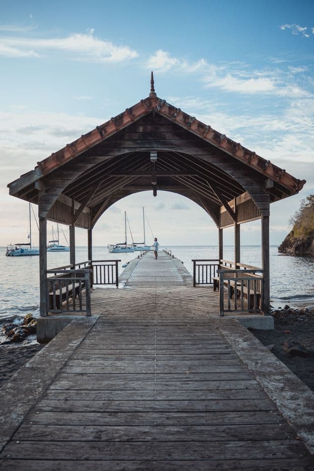 Una persona è in piedi alla fine di un lungo molo di legno, incorniciata da un arco coperto, con barche a vela sull'acqua.