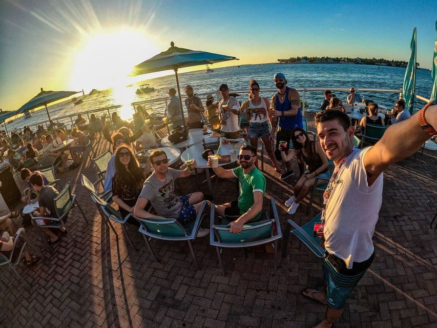 Un hombre se toma una selfie gran angular de su viaje en grupo de WeRoad en un bar concurrido frente al mar durante un atardecer vibrante.