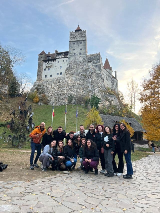 Un gruppo WeRoad in viaggio posa per una foto su un sentiero acciottolato davanti a un castello medievale su una collina rocciosa.