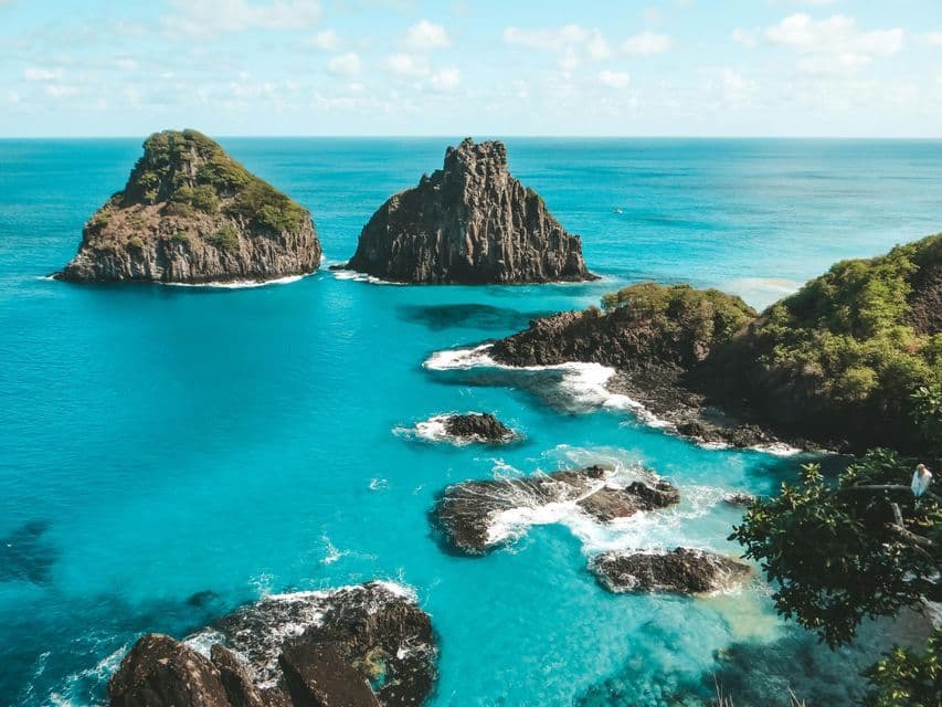 Due grandi formazioni rocciose dalla cima verde si ergono in un mare turchese, accanto a una costa frastagliata, sotto un cielo azzurro.
