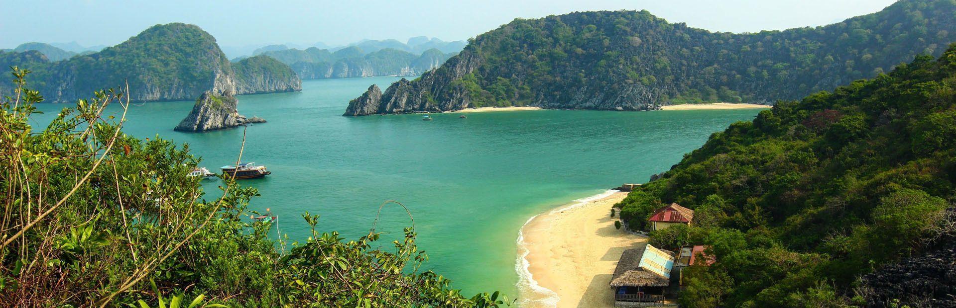 Ein Panoramablick auf eine tropische Bucht mit türkisfarbenem Wasser, üppig grünen Karstfelsen und einem kleinen Sandstrand mit Hütten.