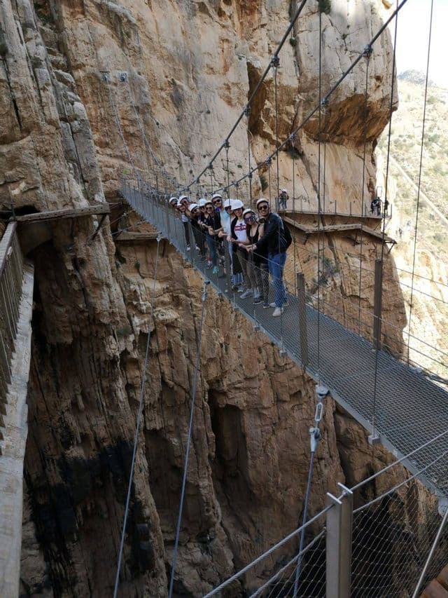 Un gruppo WeRoad in viaggio, con i caschi, posa per una foto su un ponte sospeso attaccato a una parete rocciosa a picco.