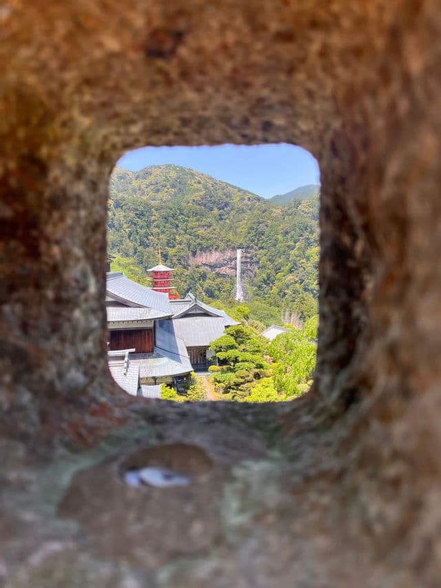 Un tempio giapponese e una pagoda rossa incorniciati da un'apertura in pietra, con una cascata che scende da una montagna verde lontana.