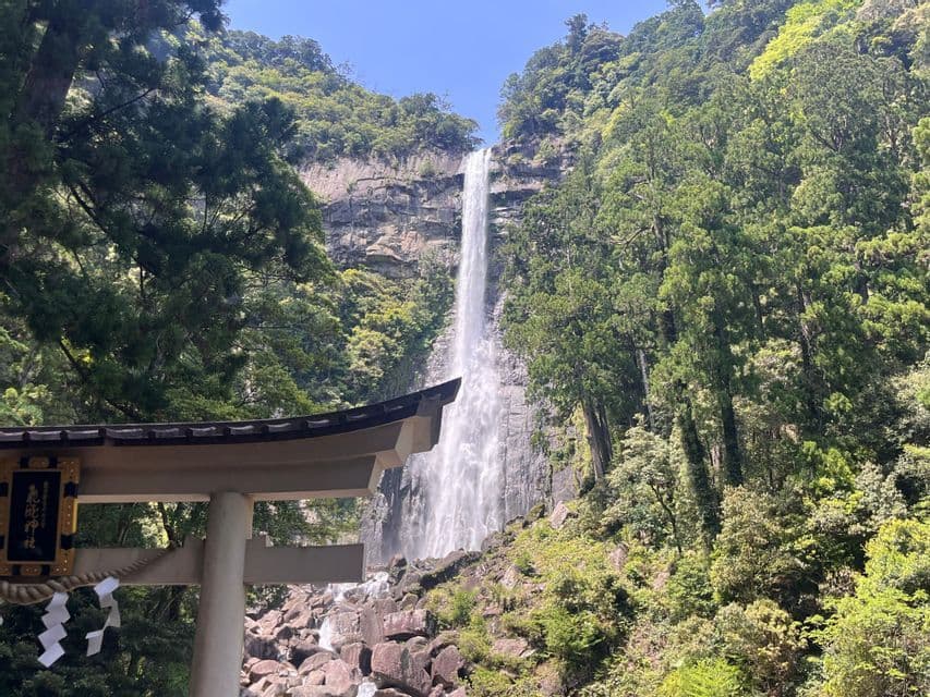 Una cascata imponente scende da una scogliera rocciosa in una fitta foresta, vista da dietro il tetto di un tradizionale torii giapponese.