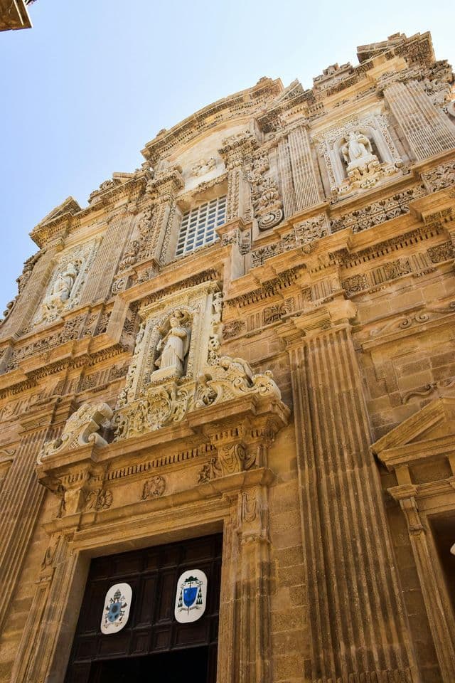 Un'inquadratura dal basso di una facciata barocca riccamente ornata di un edificio, con intagli in pietra e statue sotto un cielo sereno.