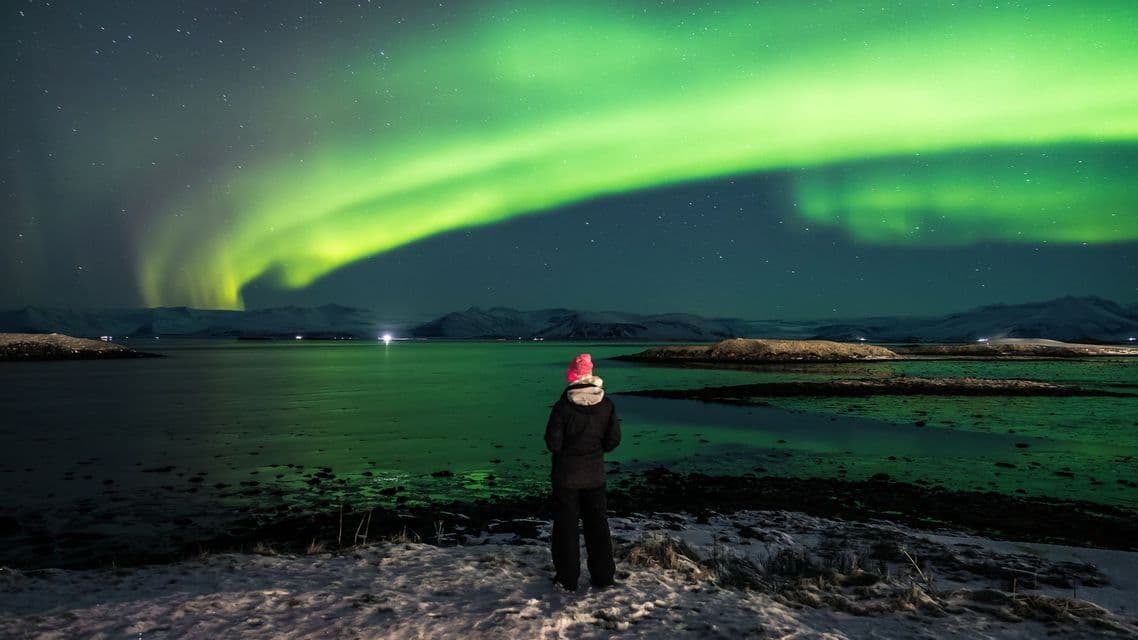 Una persona vista di spalle si erge su una riva innevata, ammirando l'aurora boreale verde su un lago e montagne distanti.
