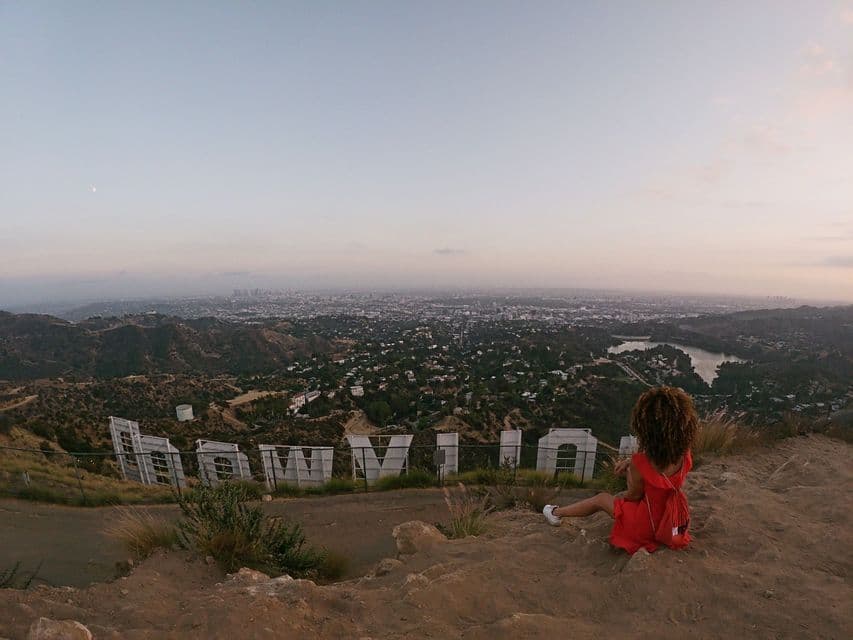Una mujer con un vestido rojo se sienta en una colina detrás del letrero de Hollywood, contemplando una extensa ciudad al anochecer.