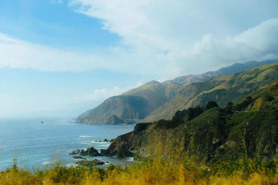 A view of a rugged coastline where green mountains slope down to the blue ocean under a partly cloudy sky, with a bridge in the distance.