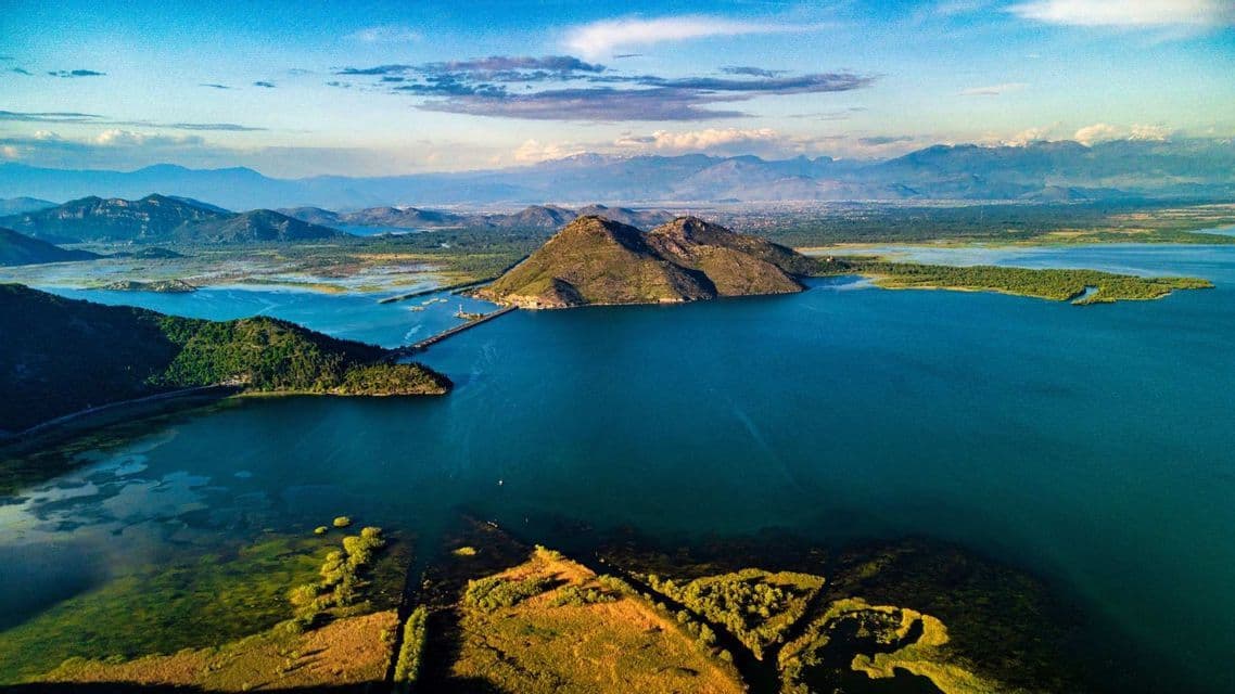 Vista aerea di un grande lago blu con un'isola rocciosa collegata alla riva da una strada rialzata, circondato da montagne.