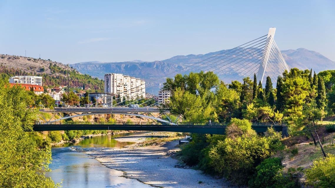 Due ponti, uno dal design moderno e strallato, attraversano un fiume in una città circondata da alberi verdi e montagne.