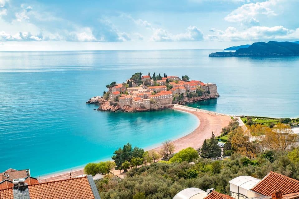 Una piccola isola fortificata con case dai tetti rossi, collegata da una strada rialzata a una spiaggia sabbiosa, circondata da un mare turchese.