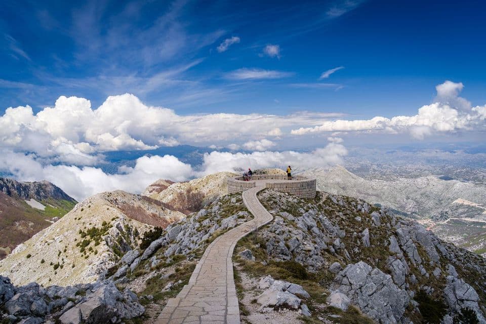 Un viaggio di gruppo WeRoad su un punto panoramico circolare in pietra, in cima a una montagna, con un sentiero tortuoso che sale e nuvole sotto.