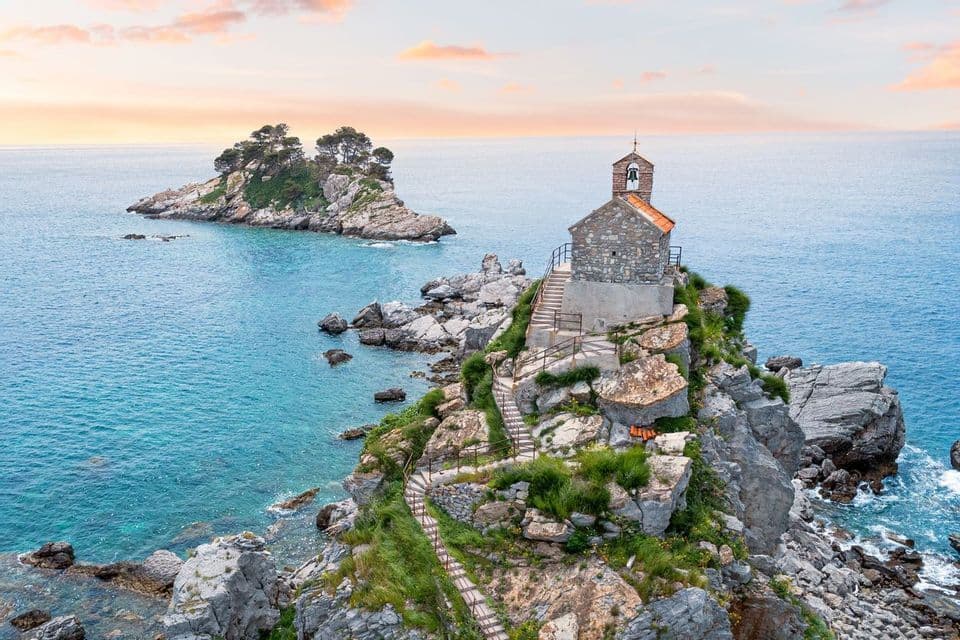Una cappella in pietra si trova su un isolotto roccioso nel mare turchese, con un sentiero tortuoso che conduce ad essa sotto un cielo al tramonto.