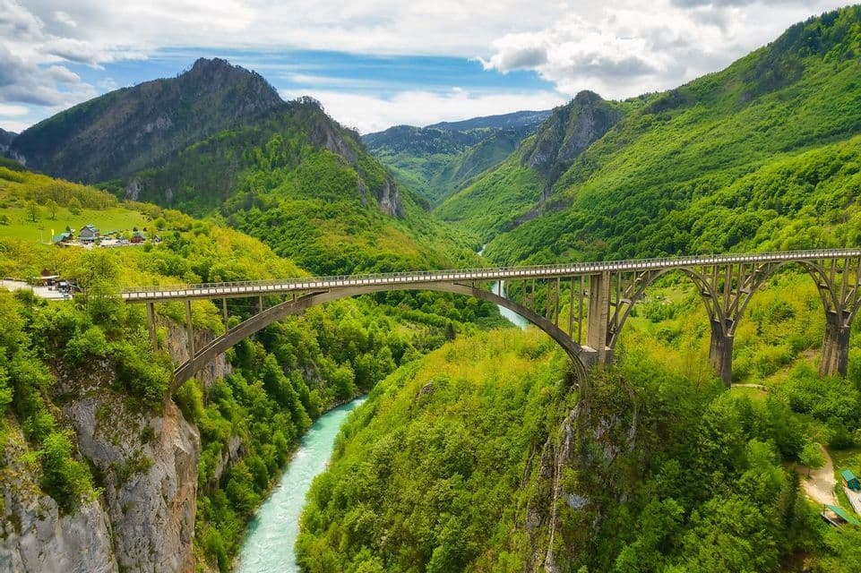 Un grande ponte ad arco in cemento attraversa un profondo canyon con un fiume turchese, circondato da montagne verdi lussureggianti sotto un cielo nuvoloso.