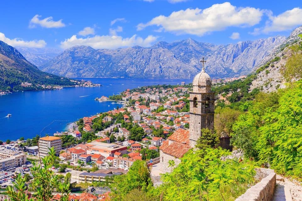 Un campanile in pietra domina una città costiera su una baia, circondata da grandi montagne sotto un cielo azzurro.