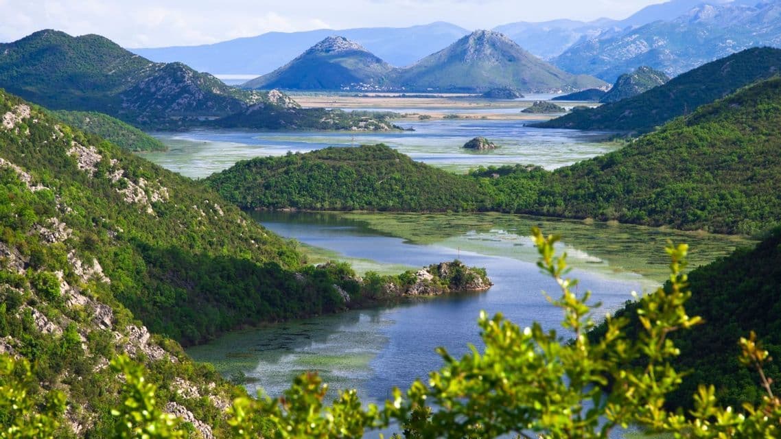 Una vista panoramica di un fiume serpeggiante con vegetazione verde, circondato da colline lussureggianti e alberate e montagne lontane.