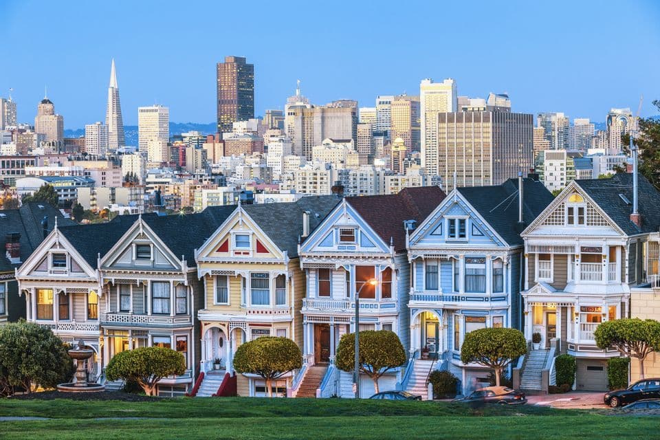 Une rangée de maisons victoriennes colorées se dressent sur une colline avec un horizon urbain moderne derrière elles sous un ciel bleu clair.