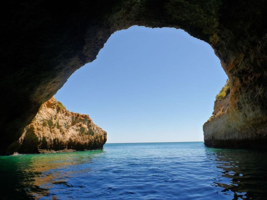 Una vista dall'interno di una buia grotta marina, che si affaccia sull'oceano azzurro illuminato dal sole e su scogliere dorate, sotto un cielo sereno.