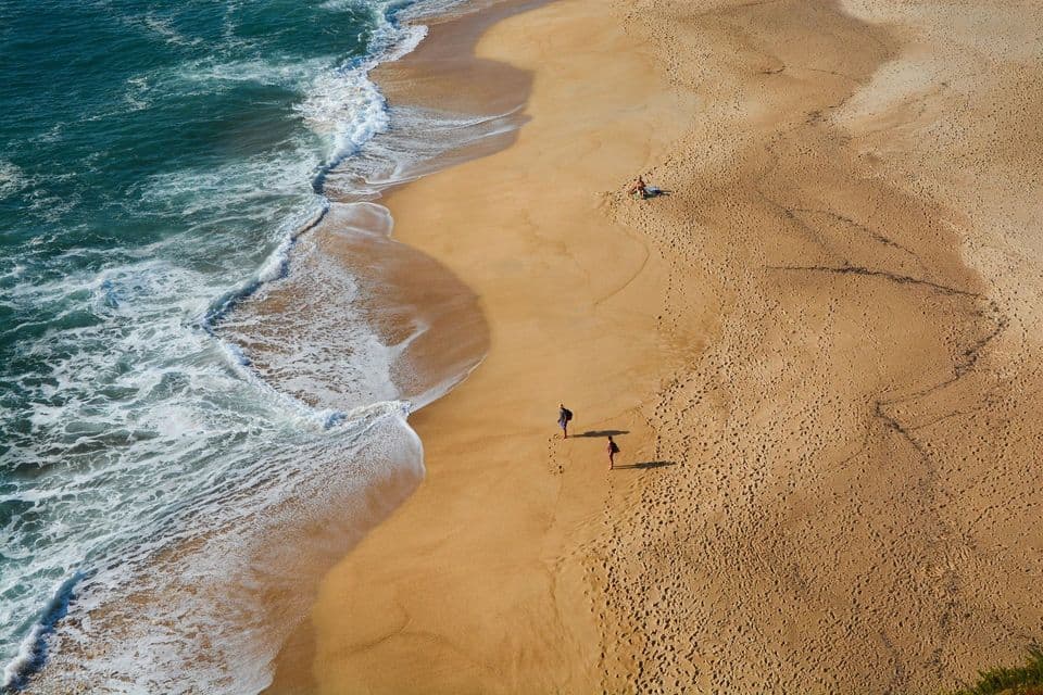 Una vista aerea di una spiaggia di sabbia dorata dove alcune persone passeggiano vicino alla riva mentre le onde turchesi si infrangono.