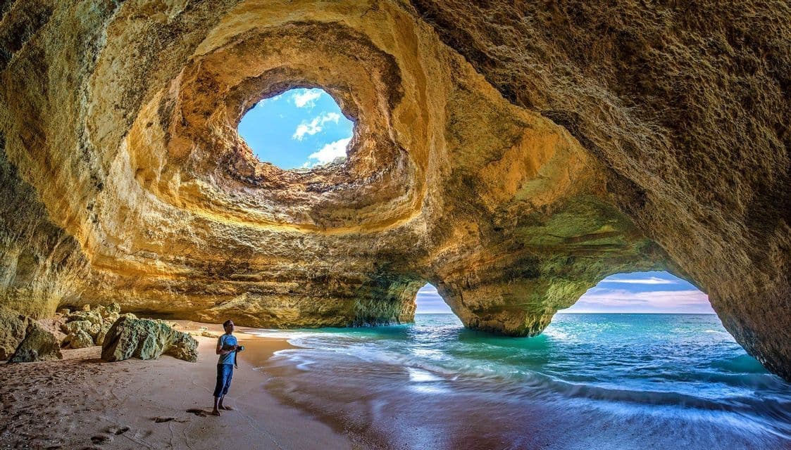 Una persona si trova su una spiaggia sabbiosa all'interno di una grande grotta marina con un'apertura circolare verso il cielo e un arco che conduce all'oceano.