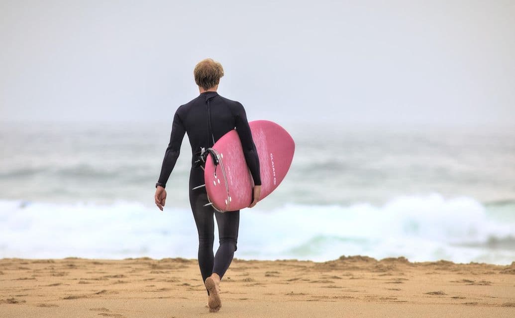 Un surfista in muta nera, visto di spalle, porta una tavola da surf rosa mentre cammina su una spiaggia sabbiosa verso l'oceano.