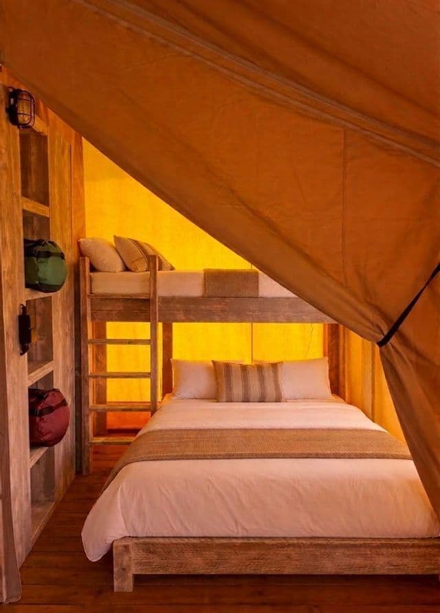 L'interno calorosamente illuminato di una tenda in tela, che mostra un letto a castello in legno posizionato sopra un letto matrimoniale, con mensole portaoggetti a lato.