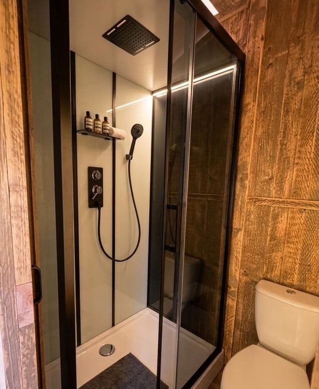 Un box doccia moderno con cornice nera e illuminazione integrata in un bagno rustico con pareti rivestite in legno.