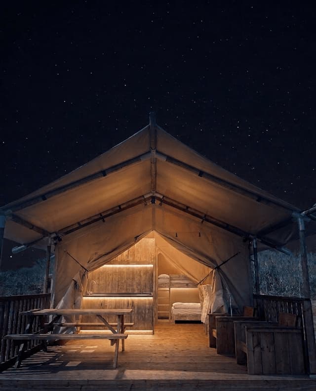 Una tenda glamping illuminata con un decking in legno e un tavolo da picnic si trova sotto un cielo notturno stellato.