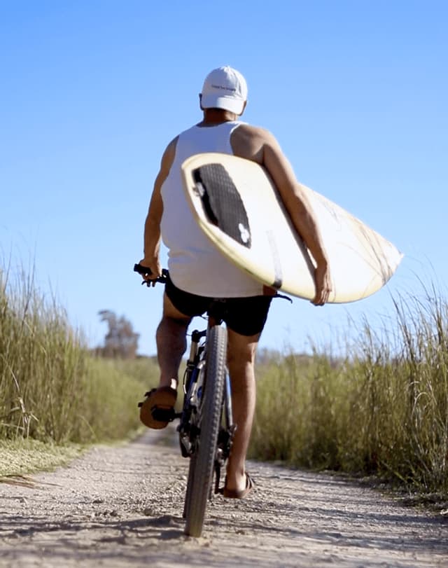 Un uomo visto di spalle va in bicicletta su un sentiero sterrato, portando una tavola da surf sotto il braccio tra erbe alte.