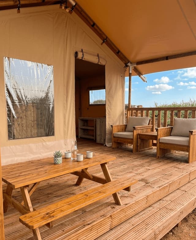 L'esterno di un'ampia tenda glamping con una terrazza in legno, un tavolo da picnic e due poltrone.